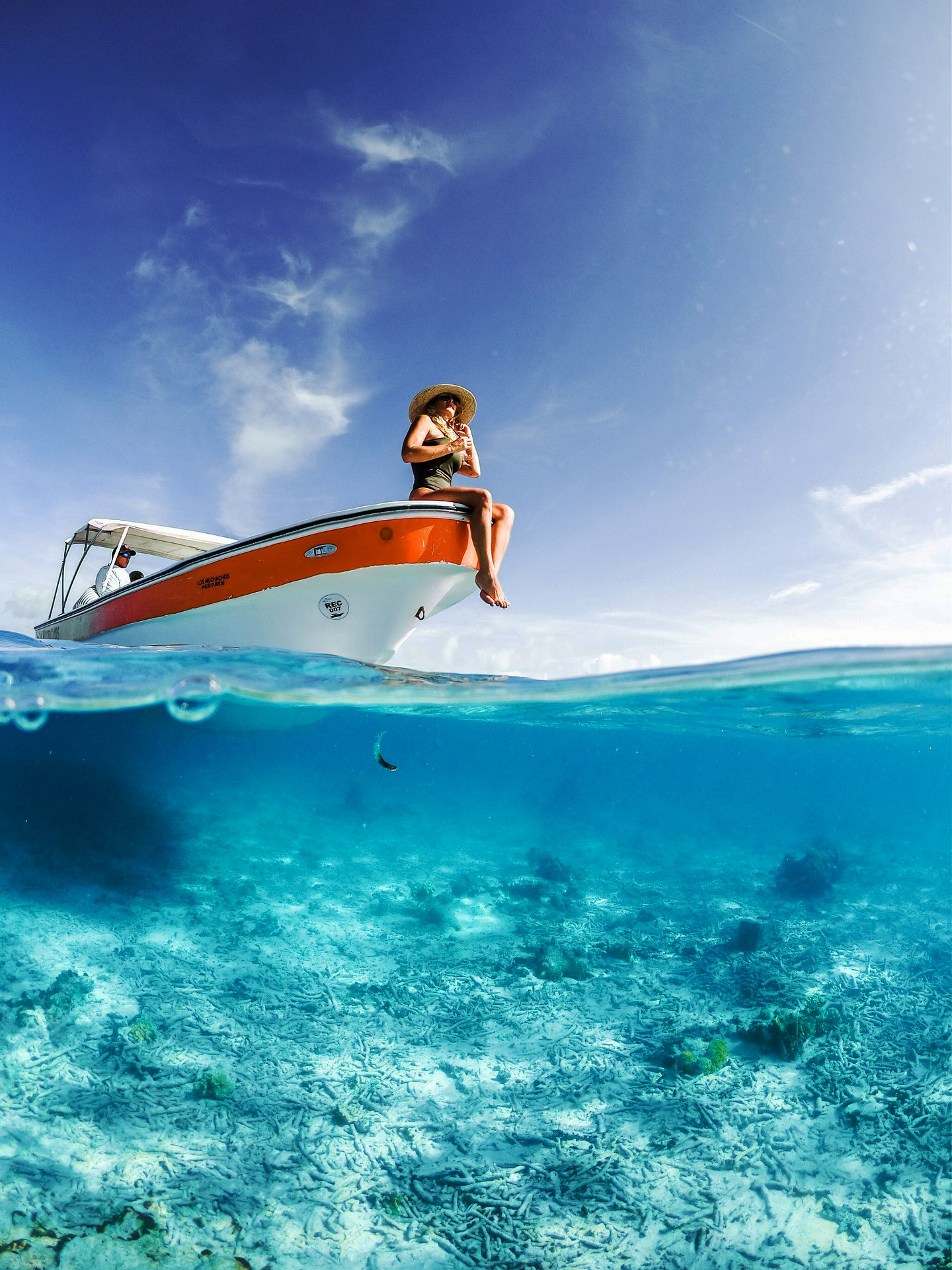 Eine Frau mit Strohhut sitzt auf der Kante eines Bootes im Meer, mit klarem Wasser und Himmel im Hintergrund.