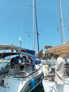 Ein Segelboot im Hafen, umgeben von anderen Booten, mit hoch aufragendem Mast und blauen Sonnensegeln.